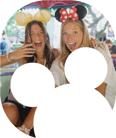 Dos jóvenes sonriendo y usando orejas de Minnie en un parque temático de Disney, detrás de una silueta decorativa.